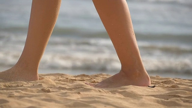 近距离的女性的脚走在金色的沙滩与海浪的背景。年轻女子的双腿踩在沙子上。光着脚的女孩在海边。暑假假期。慢动作视频下载