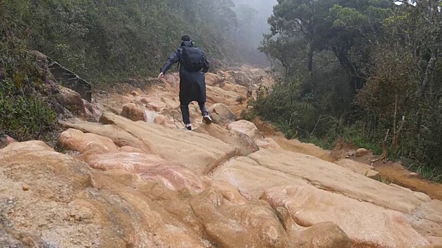年轻小伙子穿着雨衣，在岩石小道上蹦蹦跳跳。徒步旅行者背着背包在潮湿的森林里的石头地上奔跑。男游客走在丛林小径上。慢镜头后视图视频下载