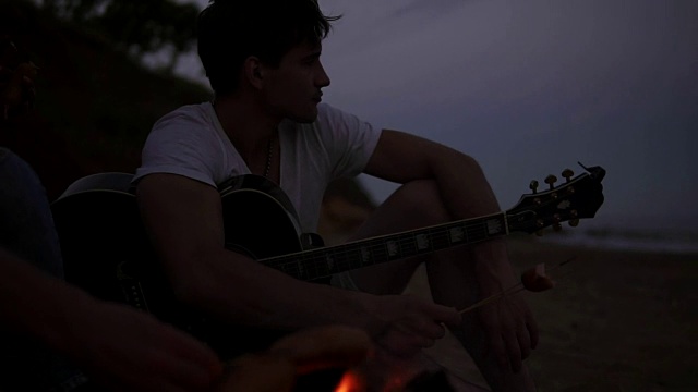 香肠在海滩火上烤的特写。晚上，一群年轻人兴高采烈地坐在海滩上的篝火旁，烤着香肠，弹着吉他。Slowmotion拍摄视频素材