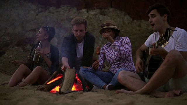 年轻人晚上在海滩上用篝火野餐。欢快的朋友们唱着歌，弹着吉他。Slowmotion拍摄视频素材