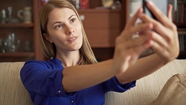 穿着蓝色衬衫的漂亮年轻女子坐在客厅的沙发上用智能手机自拍视频素材