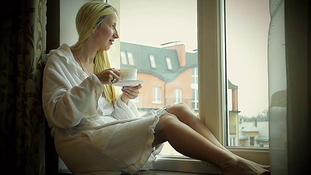 穿着浴袍的女孩在家喝咖啡，靠近窗户，看着外面视频素材