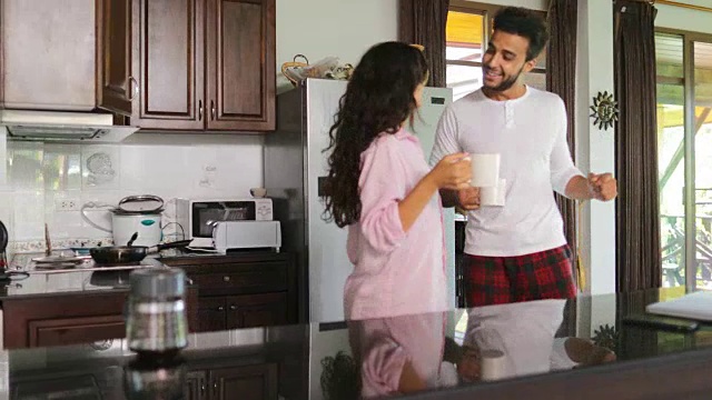 快乐的情侣早上在厨房跳舞，年轻愉快的男人和女人拿着杯子和咖啡在现代公寓工作室视频素材
