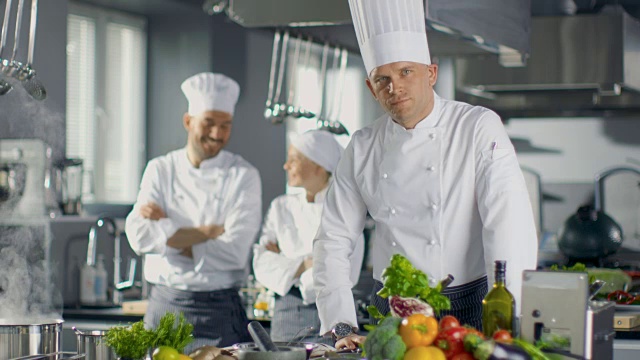 一家大餐厅的名厨准备菜肴，在镜头前微笑。背景中的两个学徒和现代厨房。视频素材
