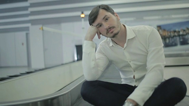 一个无聊的年轻人坐在机场行李传送带前视频素材