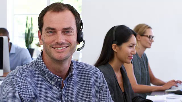 在开放式办公室里，戴着耳机的白人对着摄像头微笑视频素材