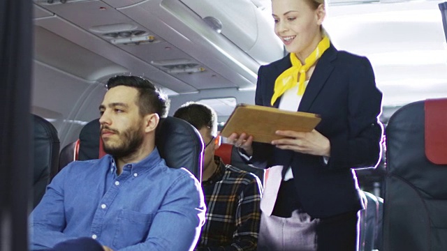 空姐向西班牙男性乘客展示带有菜单的平板电脑。他们在机上。商务舱的一个商业航空内部可见。视频购买