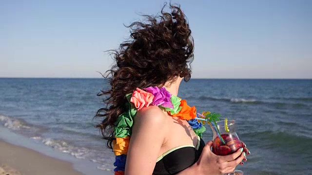 慢镜头，风中摇曳着美丽的卷发的女孩在海边拿着鸡尾酒玩着相机，性感的年轻女子视频素材