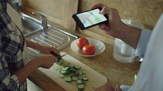 一对迷人的情侣在厨房做饭，然后用智能手机拍照在家分享社交媒体视频素材
