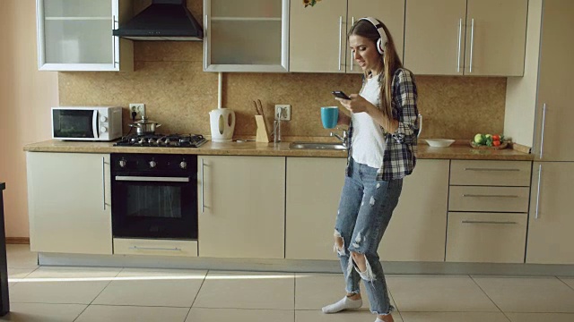 迷人的年轻有趣的女人戴着耳机在厨房跳舞和唱歌，在家里的早上和有乐趣的假期视频素材