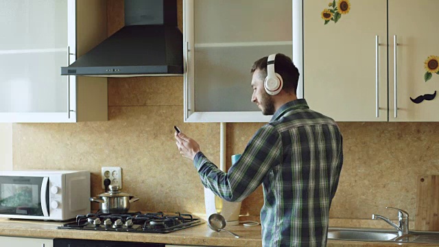 英俊的年轻有趣的男人戴着耳机在厨房跳舞和唱歌，在家里的早上和有乐趣的假期视频素材