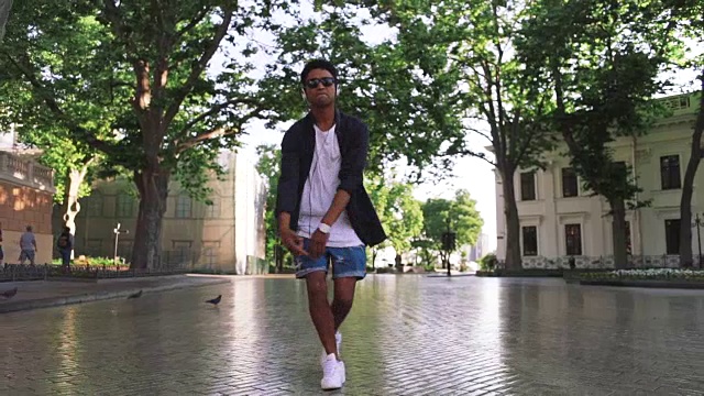 年轻时髦的黑人男子戴着白色耳机和太阳眼镜，在市中心的户外跳舞，玩一些有趣的慢动作视频素材