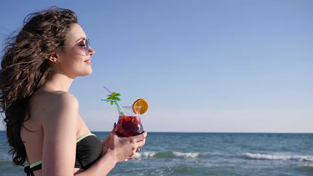 性感的女人与卷发饮料多彩的鸡尾酒，微笑的女孩在海滩上的肖像，闪亮的海背景视频素材