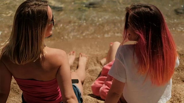 两个女人坐在沙滩上，摇晃着直发，玩得很开心视频素材