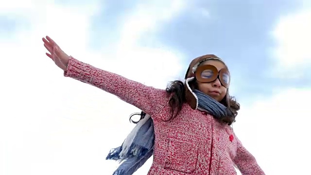 戴着飞行员帽的小女孩像一架飞机在蓝天下伸展双臂视频素材