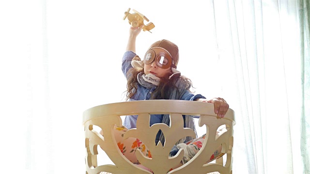 戴着飞行员帽的小女孩在公寓的椅子上玩木制飞机，4к视频下载