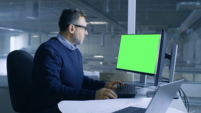 首席男性工程师在他的个人电脑上工作的技术项目。第二显示显示绿色屏幕模型。从办公室的窗户可以看到大工厂。视频下载