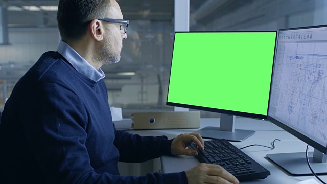 首席男性工程师在他的个人电脑上工作的技术项目。第二显示显示绿色屏幕模型。从办公室的窗户可以看到大工厂。视频素材
