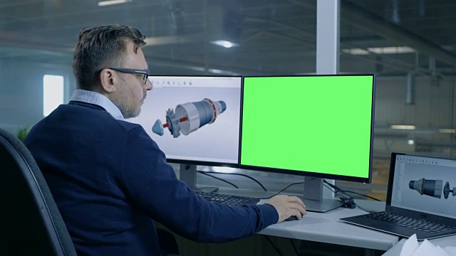 首席男性工程师为一个大的工业公司设计3D涡轮/发动机，他的第二个显示显示绿色屏幕计算机模型。从办公室的窗户可以看到大工厂。视频素材