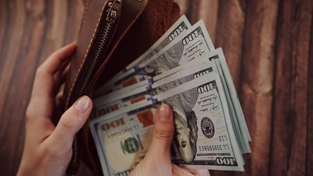 俯视图，一个女人在数，从她的棕色皮夹里拿出美元。女性拿着钱包，背景是木制的。慢动作视频下载
