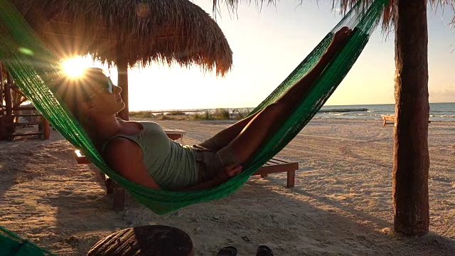 慢镜头:日落时分，年轻女子在吊床上摇摆，俯瞰沙滩视频素材