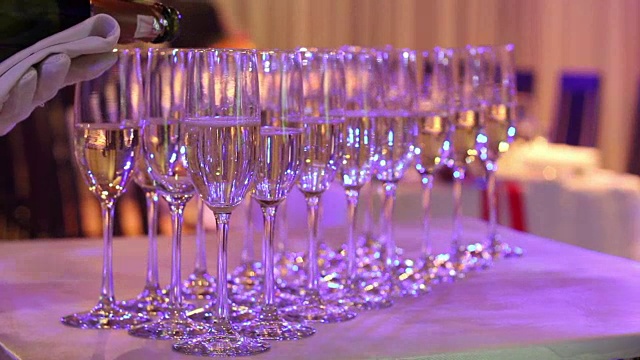 侍者将香槟倒入玻璃杯中，香槟玻璃杯放在自助餐桌上，餐厅或酒店的大厅里，侍者的手上戴着白手套拿着一瓶香槟，室内，特写视频下载