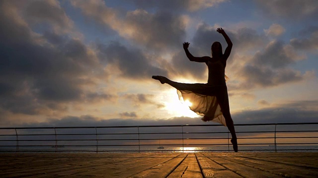 在日出或日落时，身穿米色丝绸连衣裙和尖头跳芭蕾舞者在海洋或海滩上的堤岸上。剪影的年轻美丽的金发女人长头发练习拉伸和经典的练习。史诗般的跳。慢动作视频素材