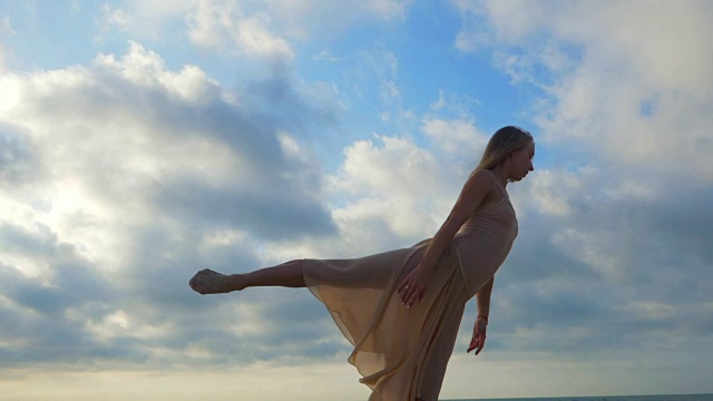 美丽的场景舞蹈芭蕾舞者在米色丝绸连衣裙和指向天空的背景。年轻美丽的金发性感女人长头发练习伸展和经典的练习。慢动作视频购买