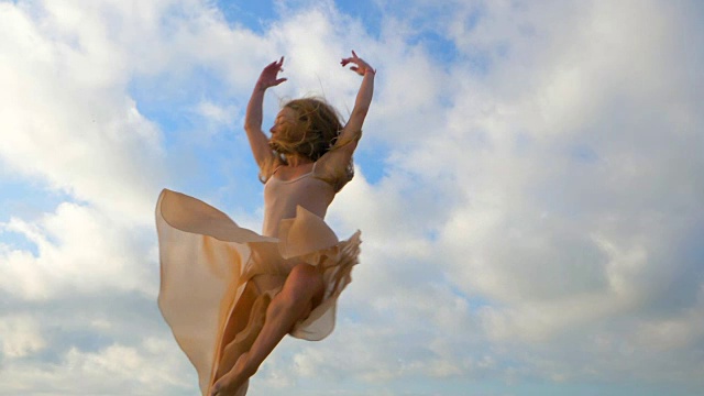 跳芭蕾舞者在米色丝绸连衣裙和指向天空的背景。剪影的年轻美丽的金发女人长头发练习拉伸和经典的练习。史诗般的跳。慢动作视频素材