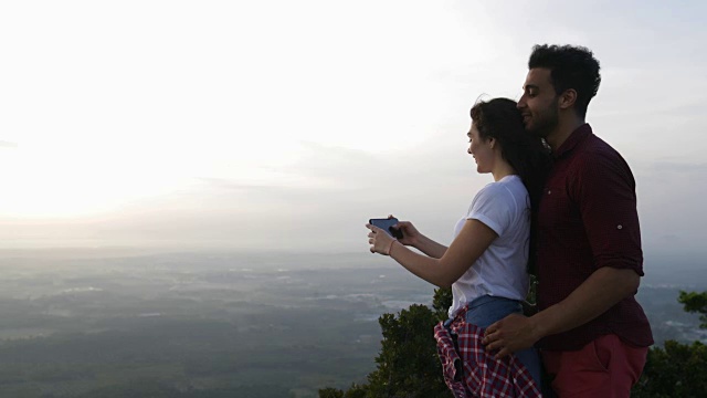 一对夫妇在山顶用手机拍摄早晨的风景，男人和女人在日出时拥抱视频素材