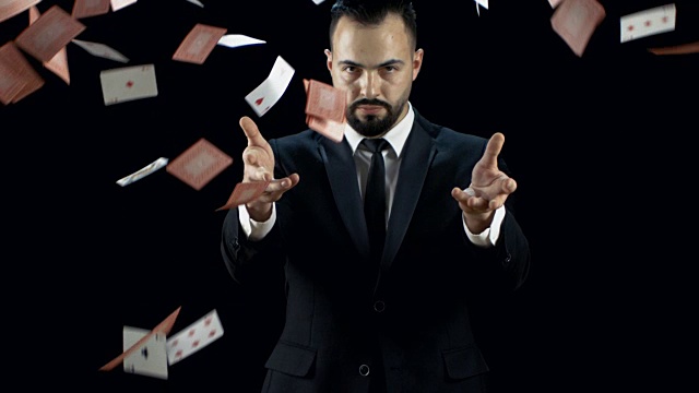 专业的年轻魔术师在一个黑色的西装扔两个喷气的纸牌到空中在慢动作。背景是黑色的。视频下载
