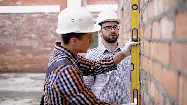 一个穿着制服的成年工程师用水平仪测量砖墙，一个戴着防护头盔的建筑师检查建筑仪器的数据视频下载