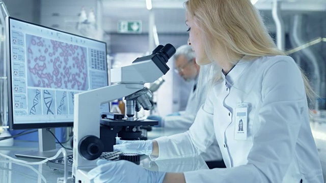 女性研究科学家在显微镜下观察生物样本。她和她的同事们在一家大型现代化实验室/医疗中心工作。视频素材