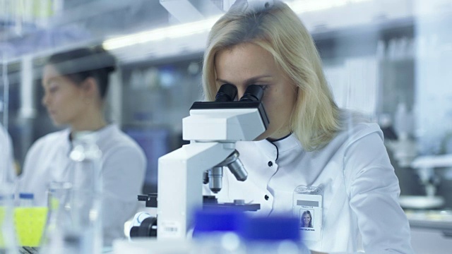 在显微镜下观察样品的女研究科学家们把滑片递给她的同事。他们在一个大型研究中心/实验室工作。视频素材