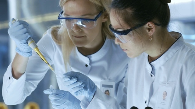 医学研究科学家把样品滴在载玻片上，她的同事在显微镜下检查它。他们在一个现代化的实验室工作。视频素材