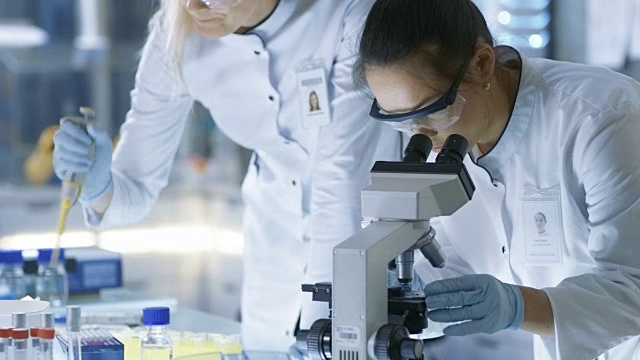 医学研究科学家把样品滴在载玻片上，她的同事在显微镜下检查它。他们在一个现代化的实验室工作。视频素材