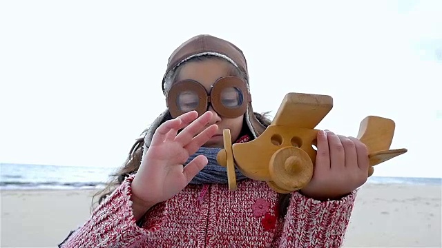 小女孩戴着飞行员帽在蓝天下玩木制飞机，慢镜头视频素材