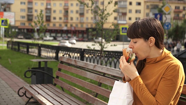 褐发女人在路边的长凳上吃汉堡包视频素材