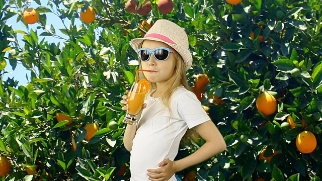 年轻的白人女孩打开和喝橙汁在花园与桔子树。夏日阳光明媚，绿油油的花园和少女站在树前。女孩戴着帽子t恤裙子和太阳镜。视频素材