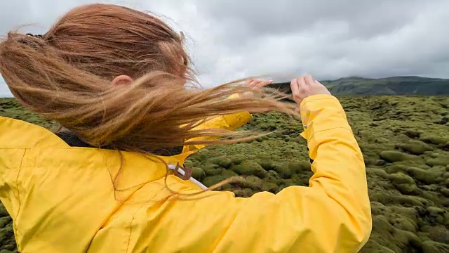 一名身穿黄色雨衣的妇女在一个多风的日子里拍摄冰岛绿色和长满苔藓的火山景观视频素材