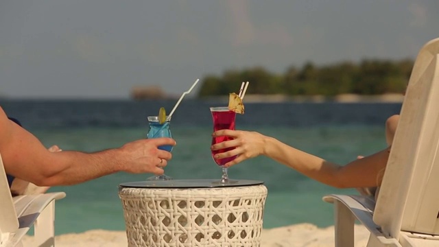 年轻的家庭夫妇日光浴和喝彩色鸡尾酒在热带海滩。情侣在蜜月异国度假躺在海边的躺椅上的阳光度假视频下载
