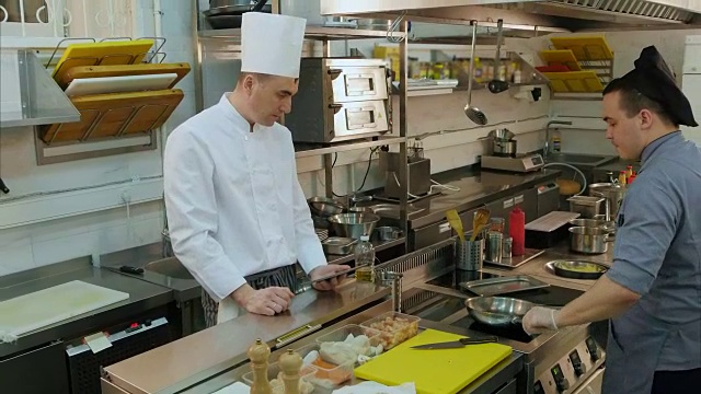 厨师用平板电脑看着他的助手在平底锅里做饭视频素材