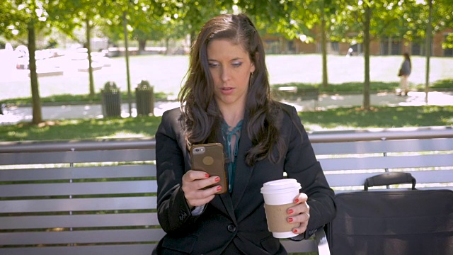 女人坐在公园的长椅上，微笑着喝着智能手机上的咖啡视频素材
