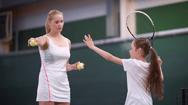 休闲区网球课的过程。女教师教年轻女运动员玩。穿着运动服的女孩，在职业球员的指导下拿着球拍击打弹跳球视频下载