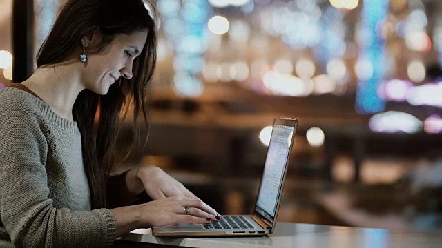 年轻漂亮的女人坐在咖啡馆靠窗的地方，正在用手提电脑。晚上在城里快乐的女性视频素材
