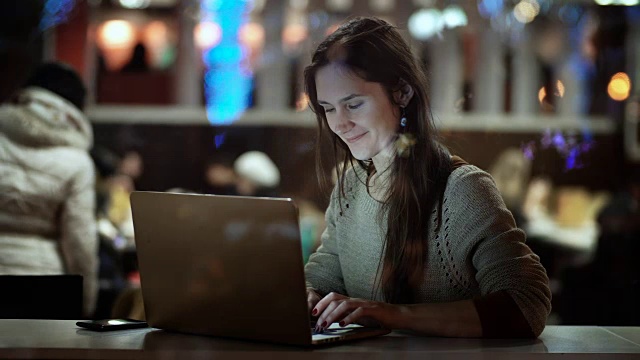 窗外的景色。年轻快乐的女人坐在晚上的咖啡馆里，用着笔记本电脑，微笑着视频素材