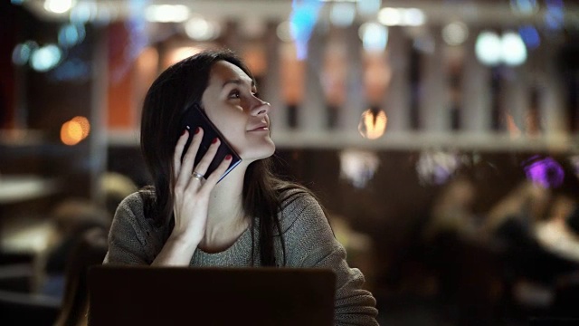 年轻漂亮的女人坐在咖啡馆里打电话。深色头发的女性在晚上使用笔记本电脑工作视频素材