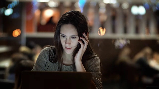 年轻漂亮的女人晚上用笔记本电脑工作。褐发女性坐在咖啡馆里用手机聊天视频素材