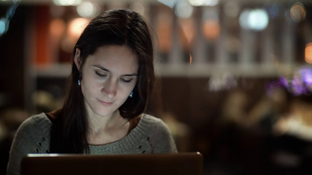 一个年轻漂亮的女人坐在咖啡馆里用笔记本电脑的肖像。晚上在城里快乐的女性视频素材
