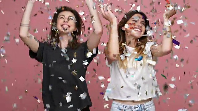 两名快乐的年轻女子在粉红色的背景上抛着闪闪发光的五彩纸屑跳舞视频下载